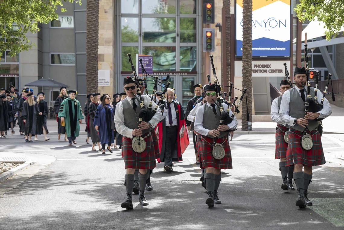 Several bagpipers in kilts and hats lead a procession of people in graduation regalia through the streets of downtown Phoenix.