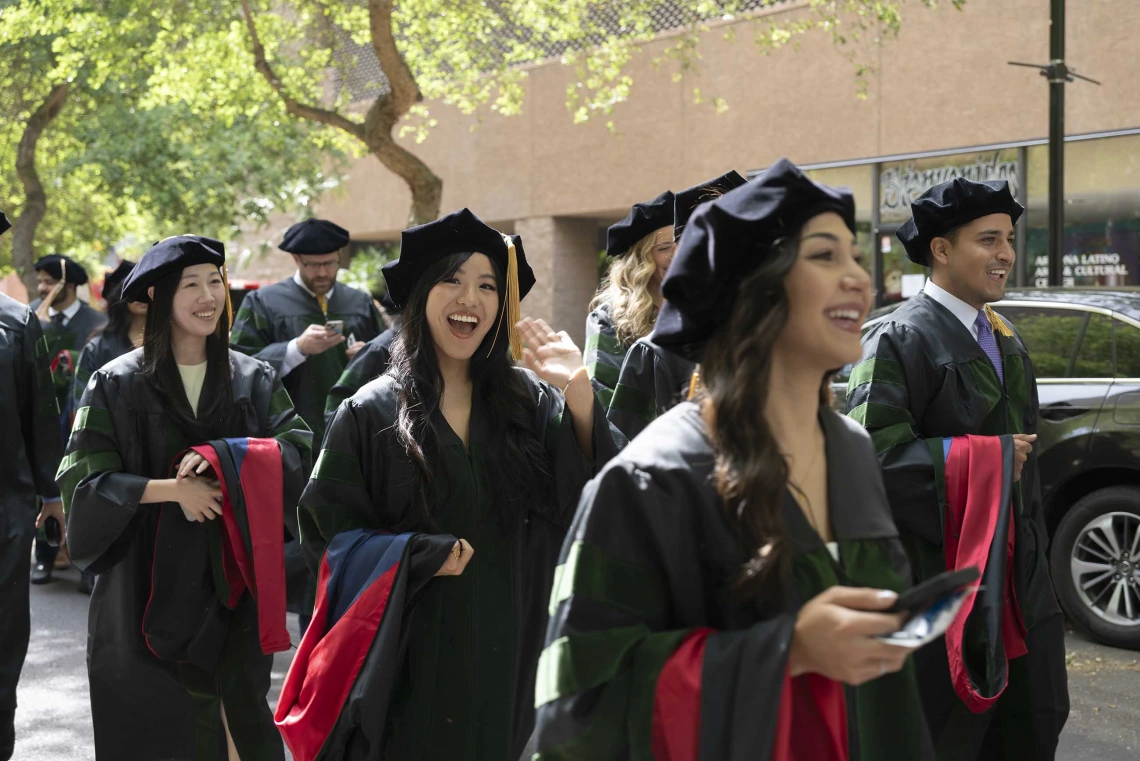 Several smiling young men and women in graduation caps and gowns walk in a line outside under some trees. 