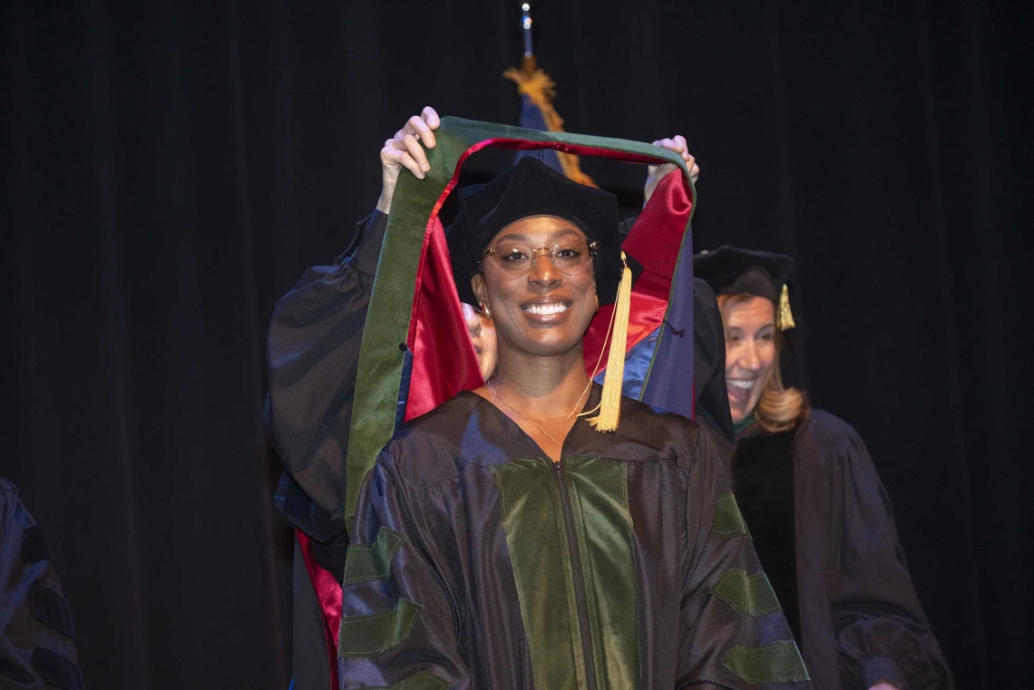 A smiling woman wearing graduation regalia looks ahead as someone behind her places a ceremonial hood over her shoulders.