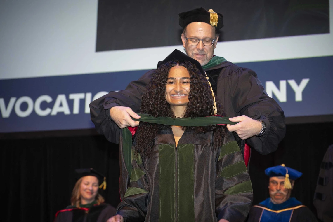 A smiling woman wearing graduation regalia looks ahead as a professor, who is also in a graduation cap and gown, places a ceremonial hood over her shoulders.