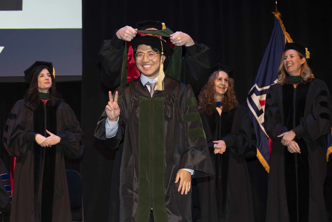 A young man wearing a graduation cap and gown smiles as he holds up two fingers in a “V” sign while a professor behind him places a ceremonial hood over his shoulders. 