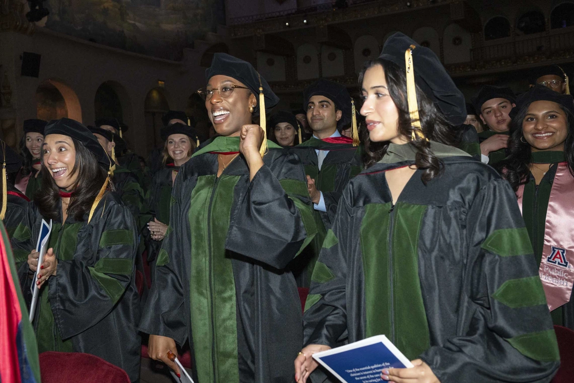 Dozens of medical students wearing graduation caps and gowns smile as they stand together during their graduation ceremony. 