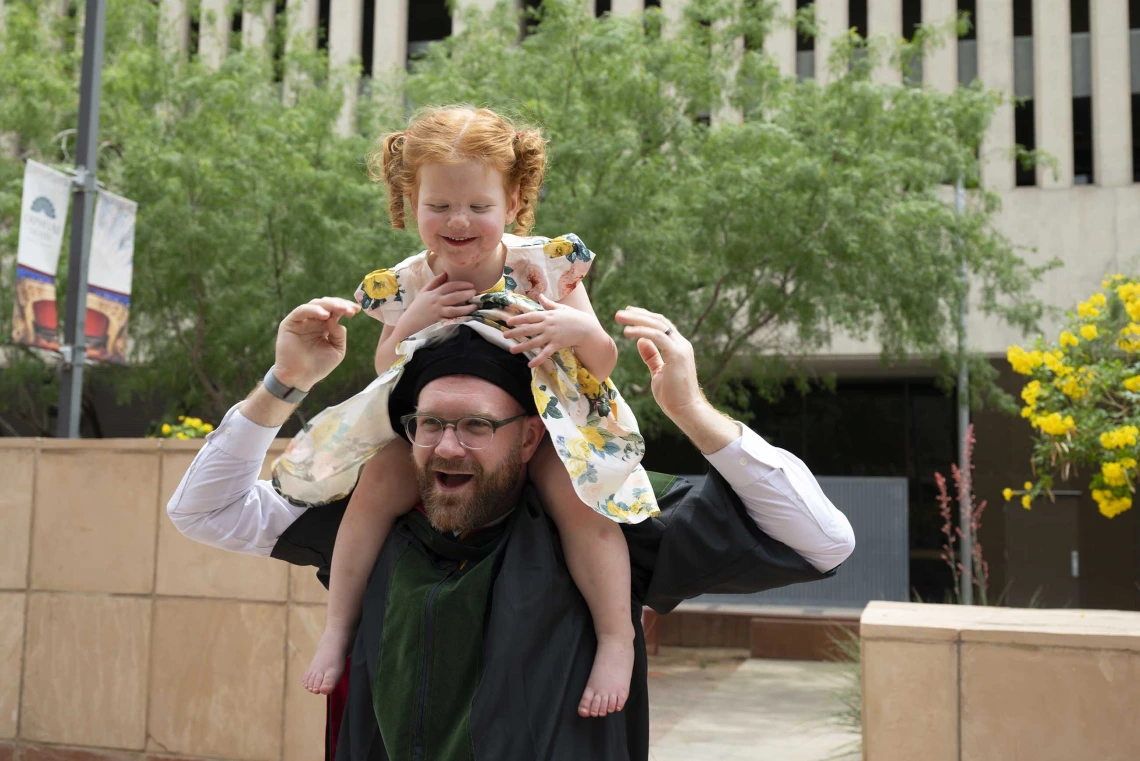 A bearded man wearing a graduation cap and gown smiles as his smiling young daughter sits on his shoulders. 