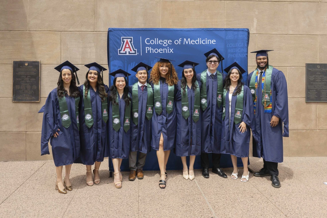 Nine students, all wearing graduation caps and gowns, stand together in front of a “College of Medicine – Phoenix” backdrop outside. 