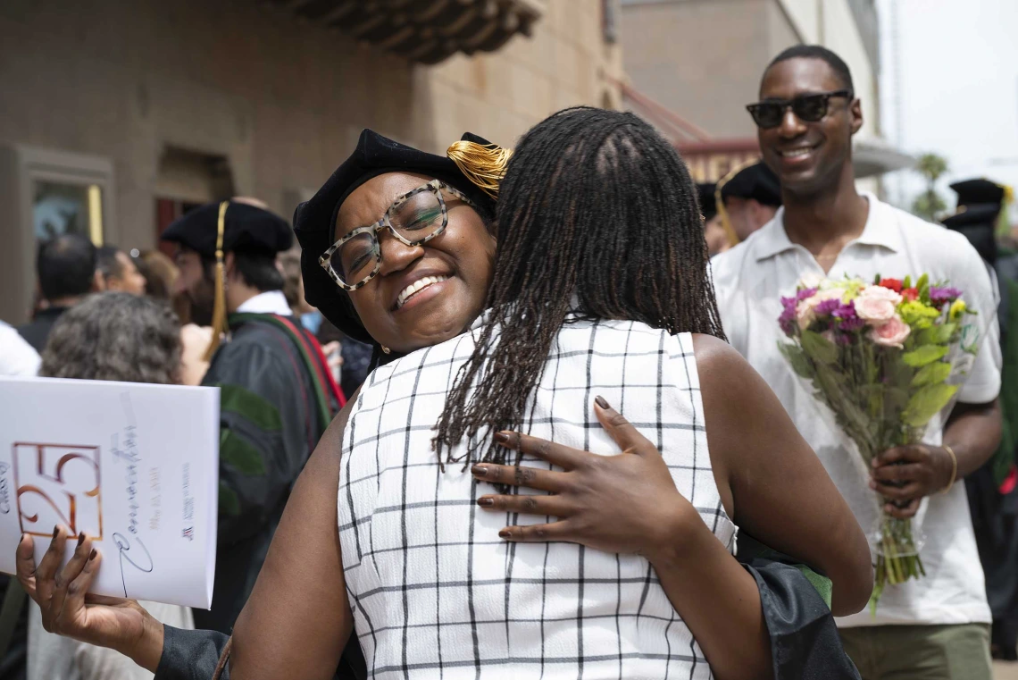 A smiling young woman in a graduation cap and gown hugs her mom after her graduation ceremony. 
