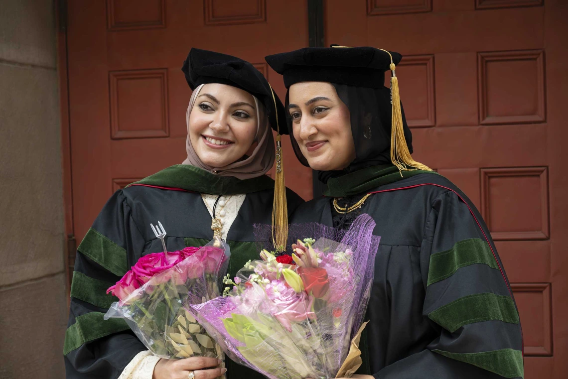 Two young women in graduation caps and gowns hold flowers and smile. 