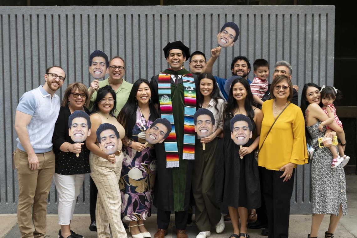 A tall, smiling young man in a graduation cap and gown stands with about a dozen family members, many of whom are holding photos of his face. 