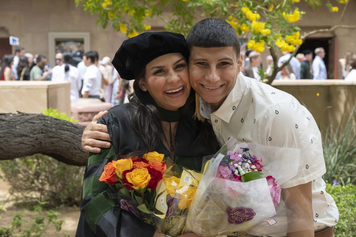 A young woman in a graduation cap and gown smiles while holding flowers and side hugging her brother. 