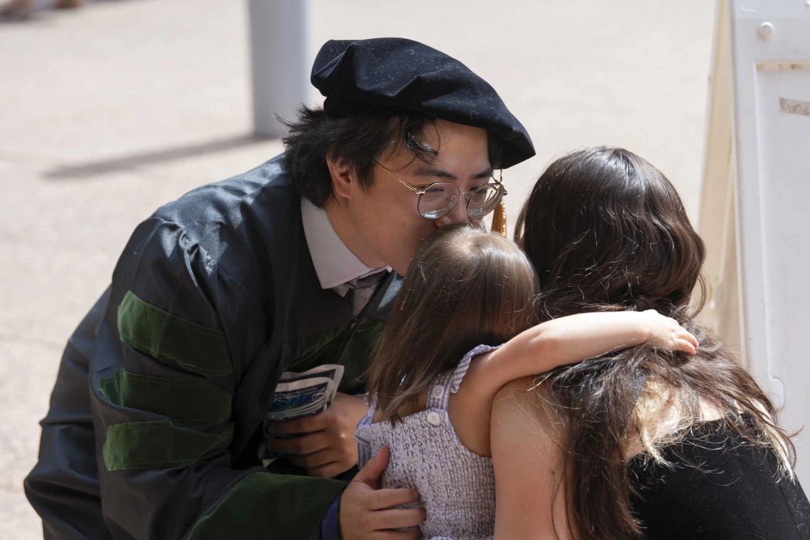 A man wearing a graduation cap and gown kisses his daughter, who is being held by his wife. 
