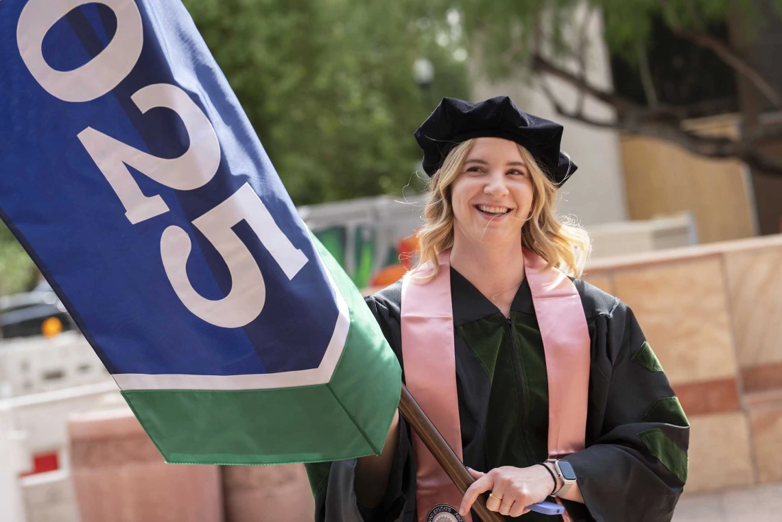 A young woman in graduation regalia smiles as she holds a large flag with “2025” on it. 