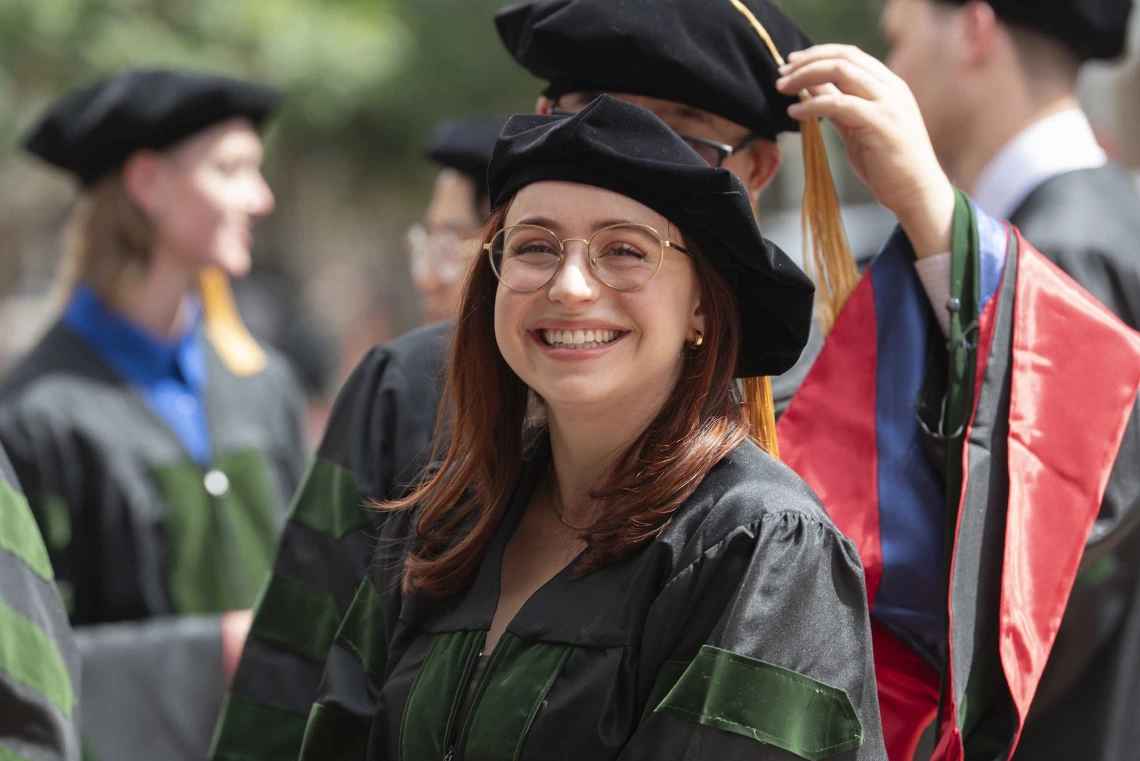 A young woman wearing graduation regalia smiles as other people dressed the same way are in the background. 