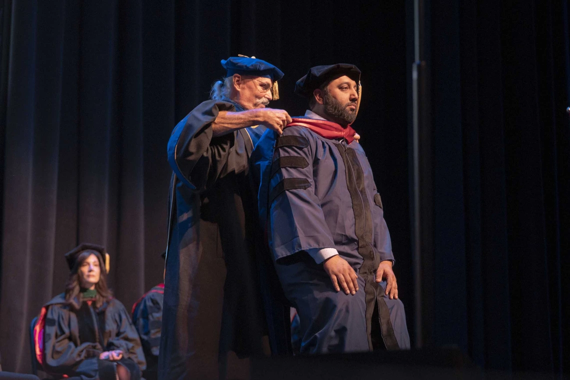 A professor places a ceremonial hood over the shoulders of a graduate onstage during a ceremony. Both are wearing graduation regalia. 