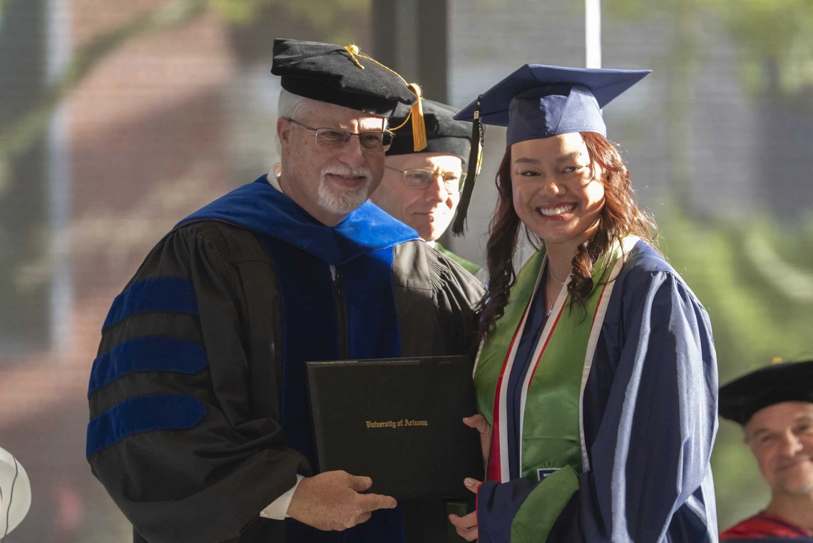 A male professor presents a diploma to a young woman. Both are smiling and wearing graduation regalia. 