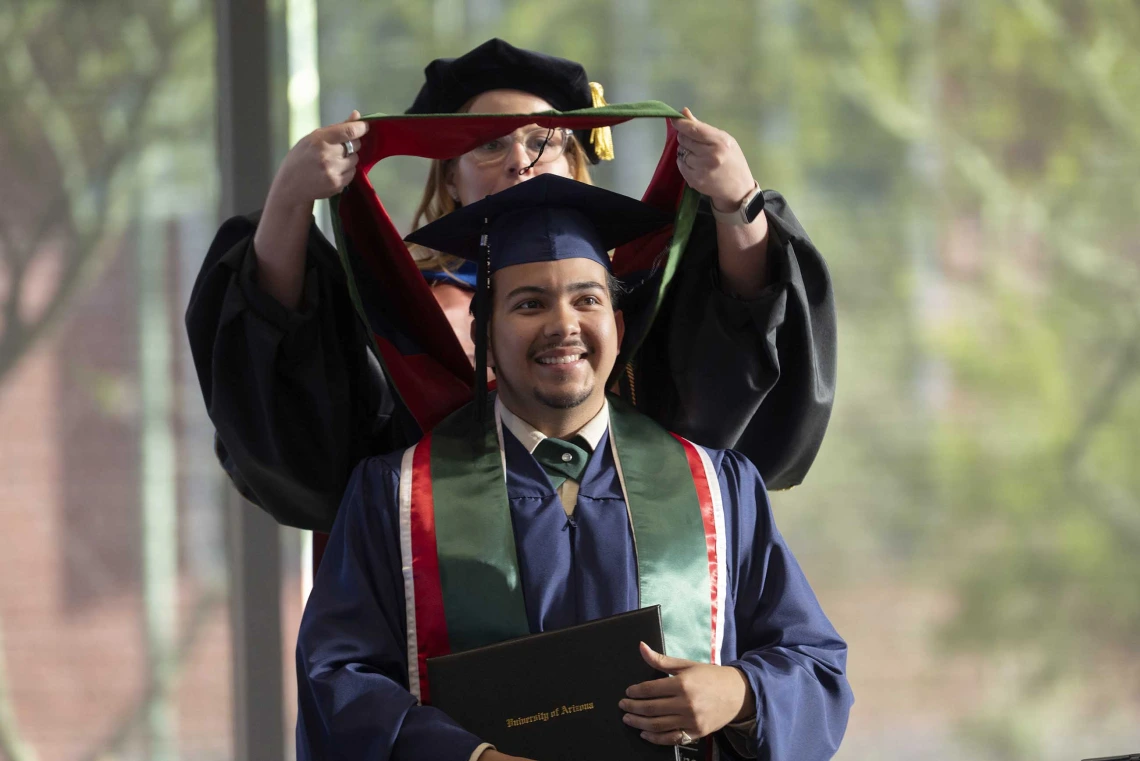 A young man wearing graduation regalia smiles as a professor standing behind him places a ceremonial hood on his shoulders.