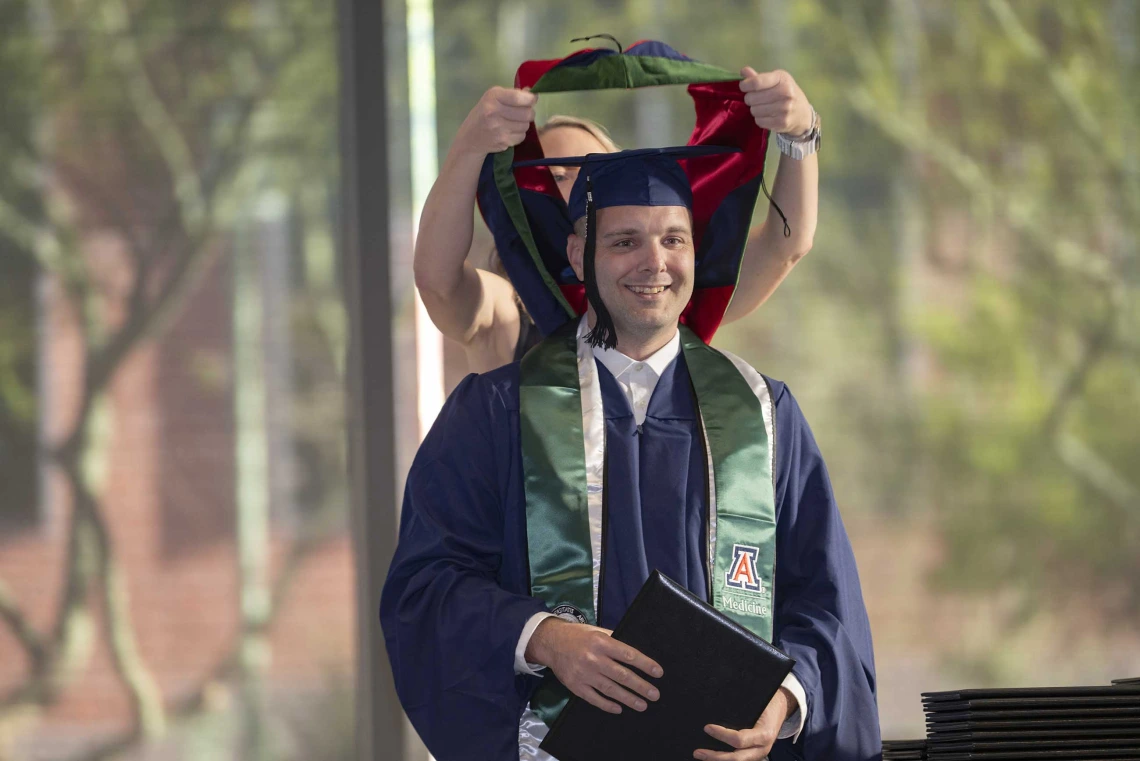 A young man in a graduation cap and gown smiles as a professor places a ceremonial hood over his shoulders. 