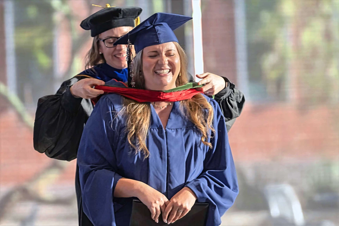 A professor places a ceremonial hood over the shoulders of a young woman. Both are smiling and wearing graduation regalia.