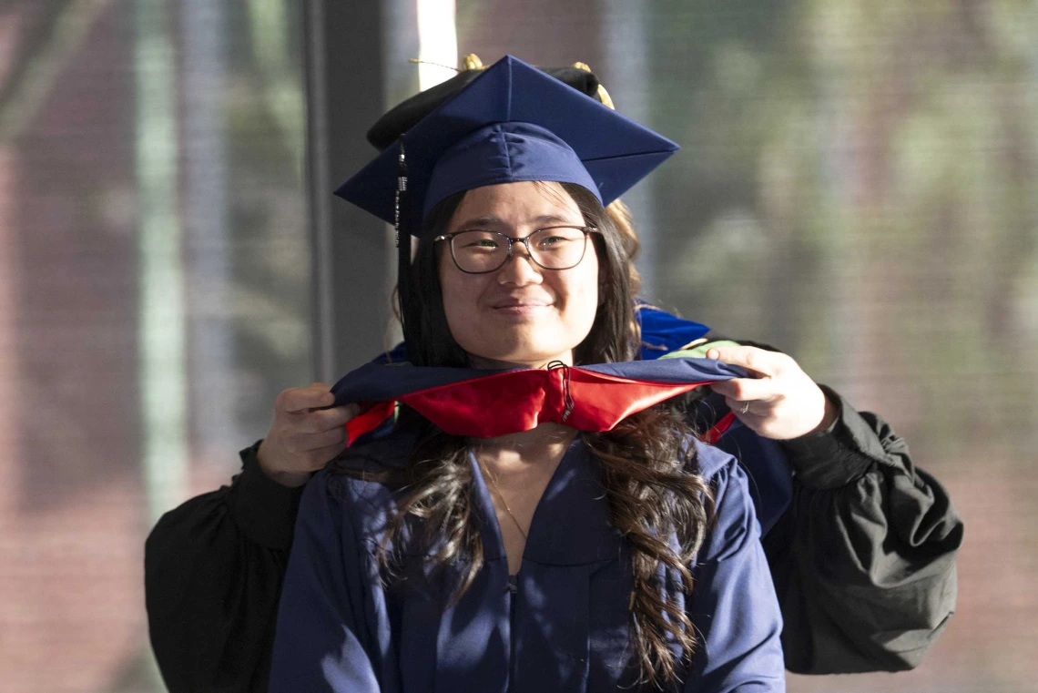 A young woman wearing graduation regalia smiles as a professor standing behind her places a ceremonial hood on her shoulders.