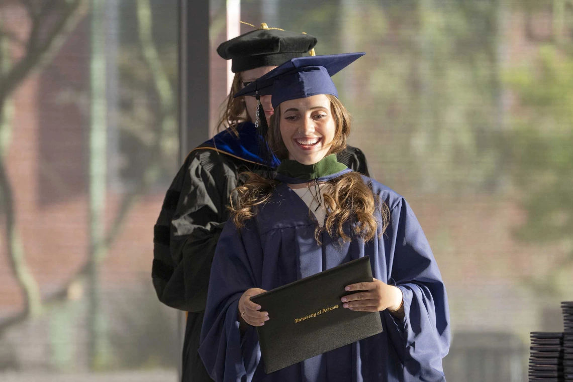 A young woman wearing graduation regalia smiles as she holds her diploma. An obscured professor stands behind her. 