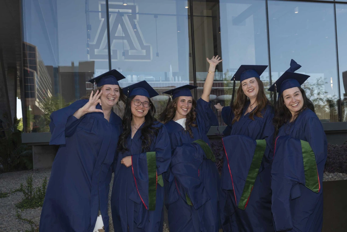 Five young women wearing graduation caps and gowns smile and cheer outside a glass building. 