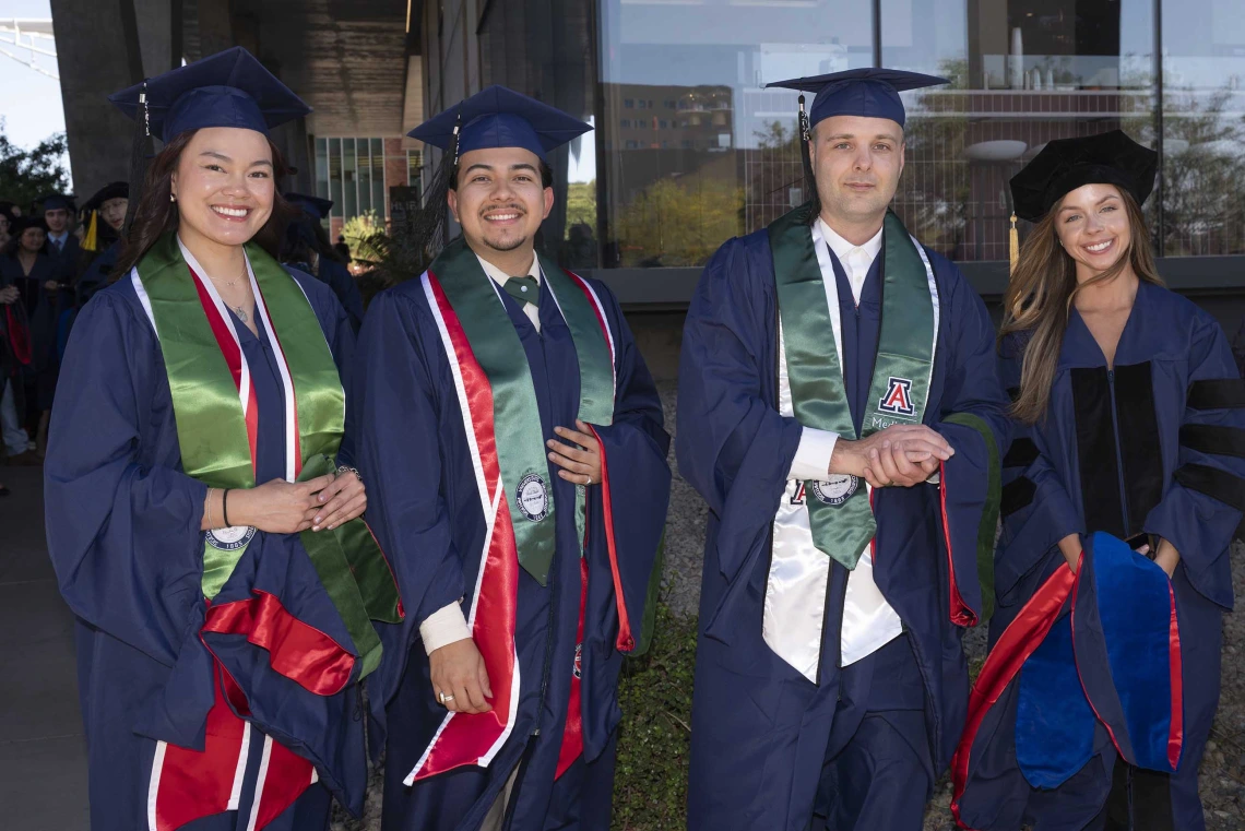 Four smiling graduates wearing caps and gowns stand outside a glass building. 