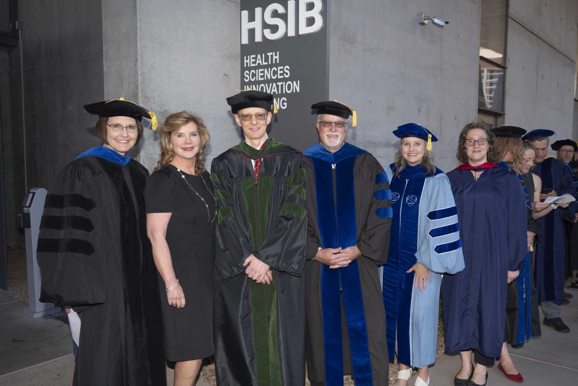 Six professors in graduation regalia stand together under a sign that reads “Health Sciences Innovation Building.”