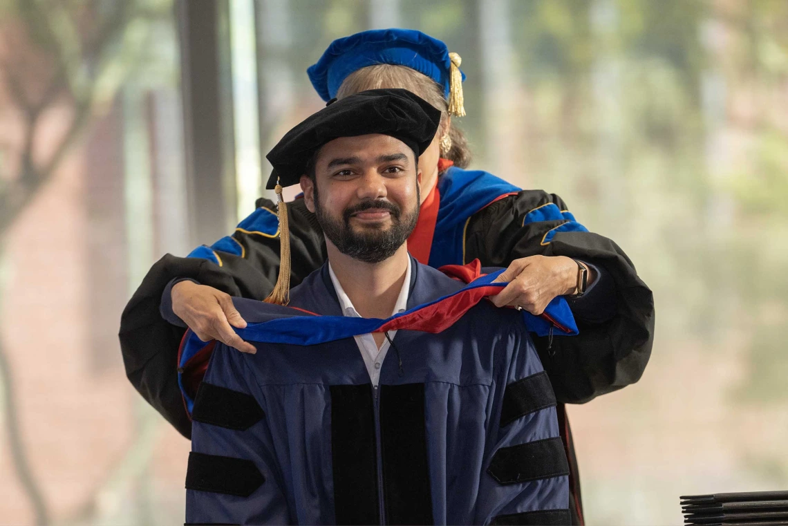 A student wearing a graduation cap and gown has a ceremonial hood placed over his shoulders by a professor. 