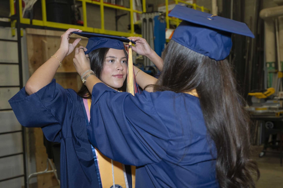 Two young women in graduation caps and gowns help each other set their mortarboards on straight. 