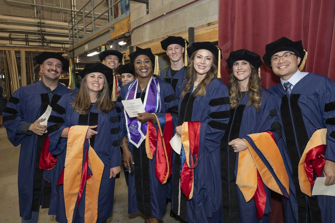Nine College of Nursing students wearing caps and gowns stand backstage waiting for their graduation ceremony to begin. 