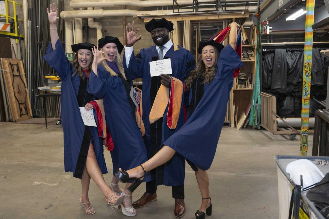 Four College of Nursing students wearing graduation caps and gowns wave and smile backstage before the graduation ceremony begins. 