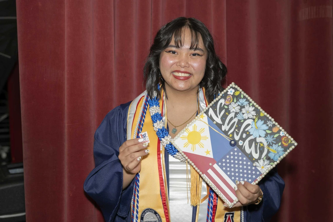 A smiling young woman wearing a graduation gown holds up her decorated mortar board and a “First Cat Grad” pin.