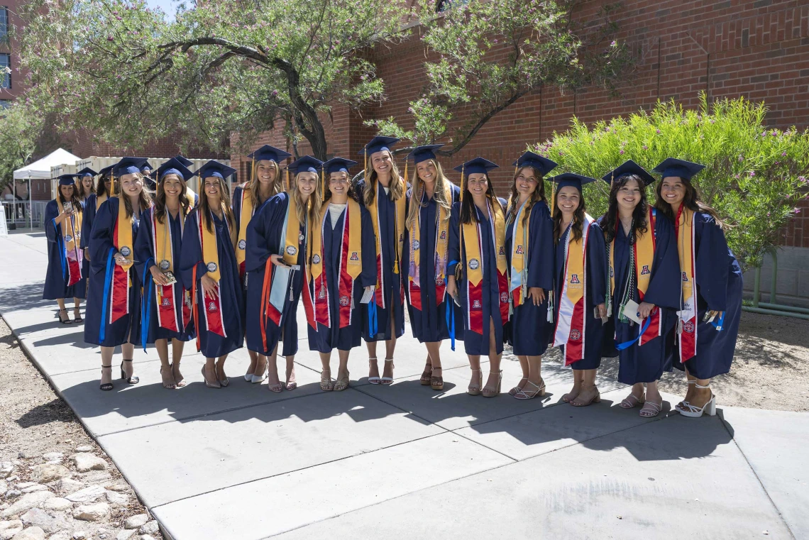 About a dozen nursing students, all dressed in graduation regalia, stand in a row outside a building. 