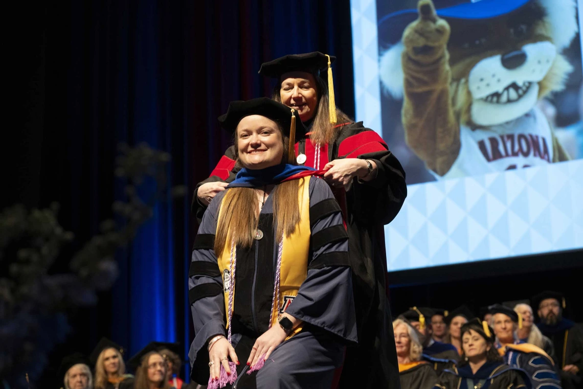A smiling young woman wearing graduation regalia has a ceremonial graduation hood placed over her shoulders by a professor.