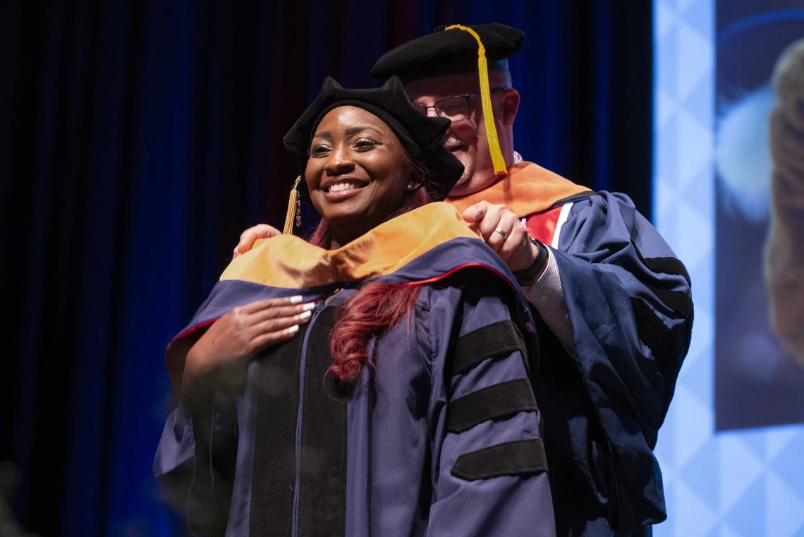 A smiling young woman wearing graduation regalia has a ceremonial graduation hood placed over her shoulders by a professor. 