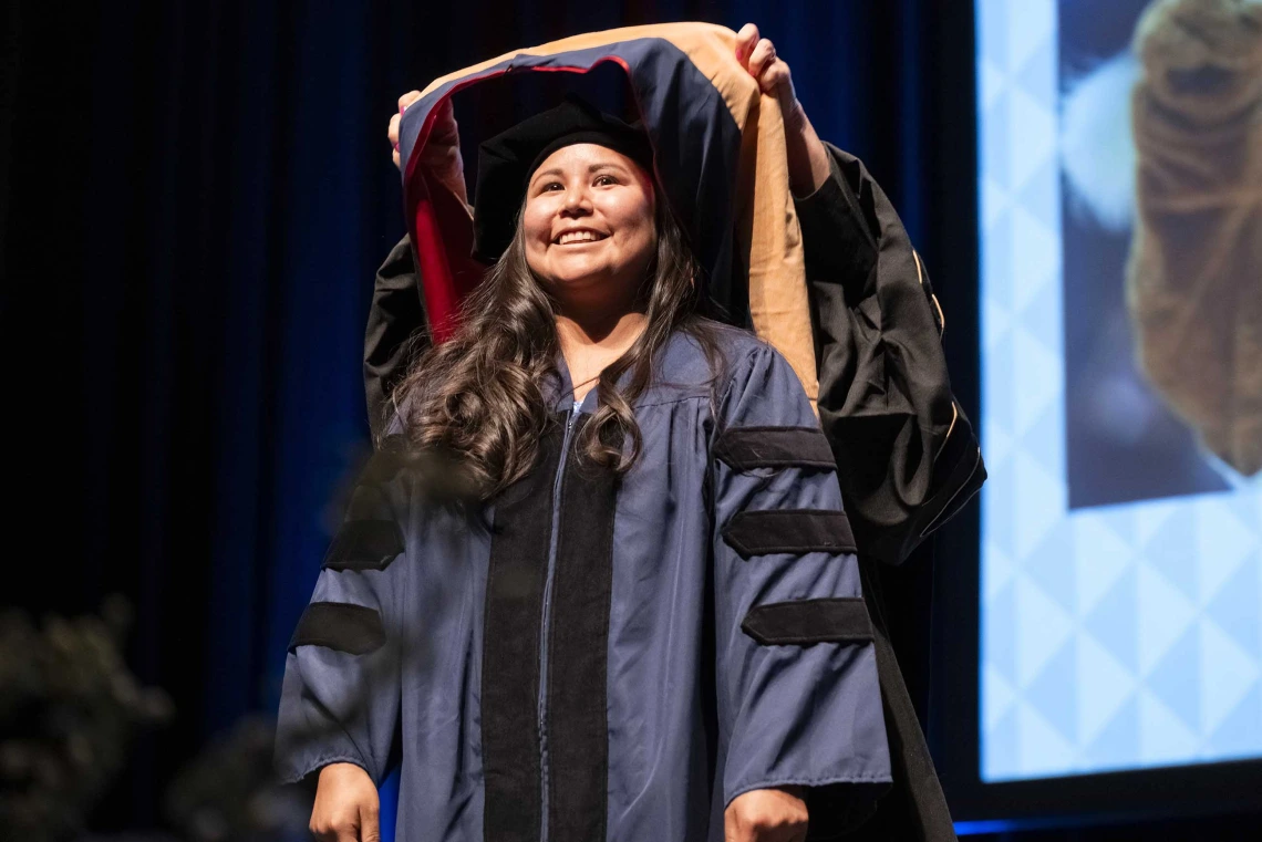 A smiling young woman wearing graduation regalia has a ceremonial graduation hood placed over her shoulders by a professor.