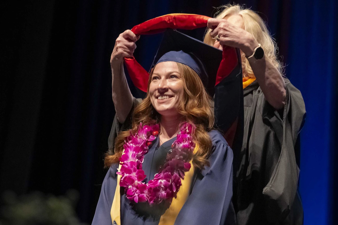 A young woman wearing graduation regalia smiles as a professor places a traditional hood over her shoulders from behind. 