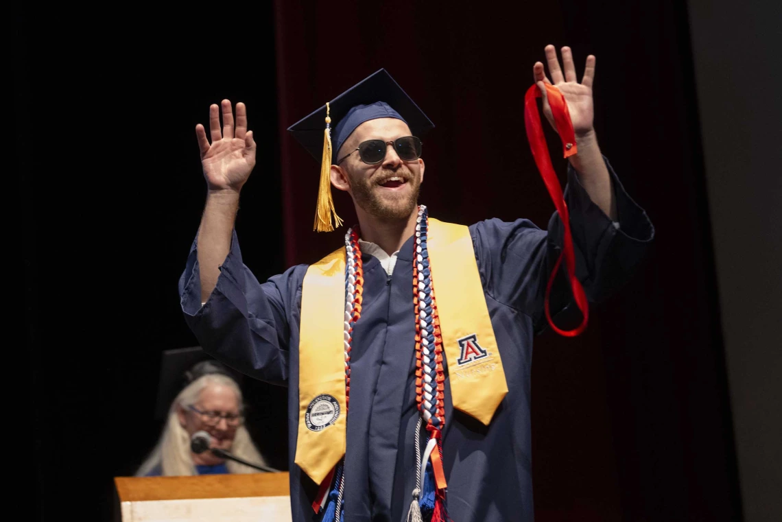 A young man wearing a graduation cap and gown and sunglasses raises his hands in the air as he walks across a stage.