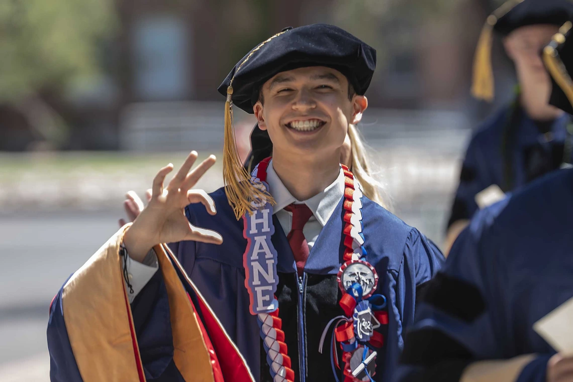 A young man in graduation regalia smiles and gives the hand sign for the University of Arizona Wildcats as he walks outside. 