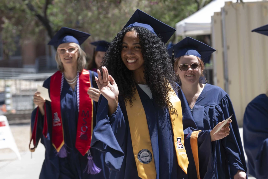 Three young women dressed in graduation caps and gowns smile as they walk in a line outside. 