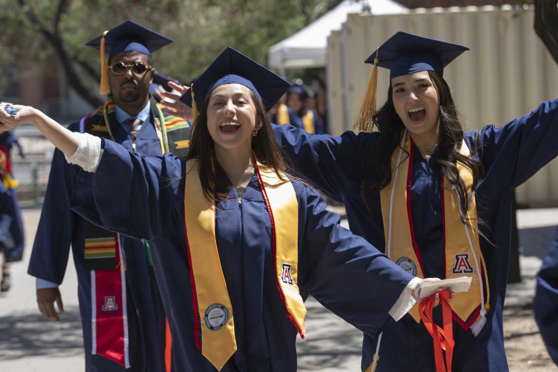 Two young women wearing graduation caps and gowns raise their hands in celebration as they walk outside with other graduates behind them. 