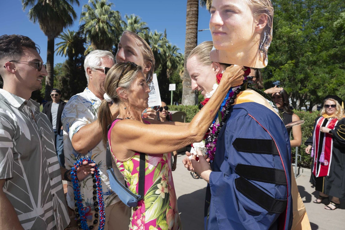 A mother places a flower lei around her son’s neck while holding a large photo of him in her hand. Her son is wearing a graduation gown. 