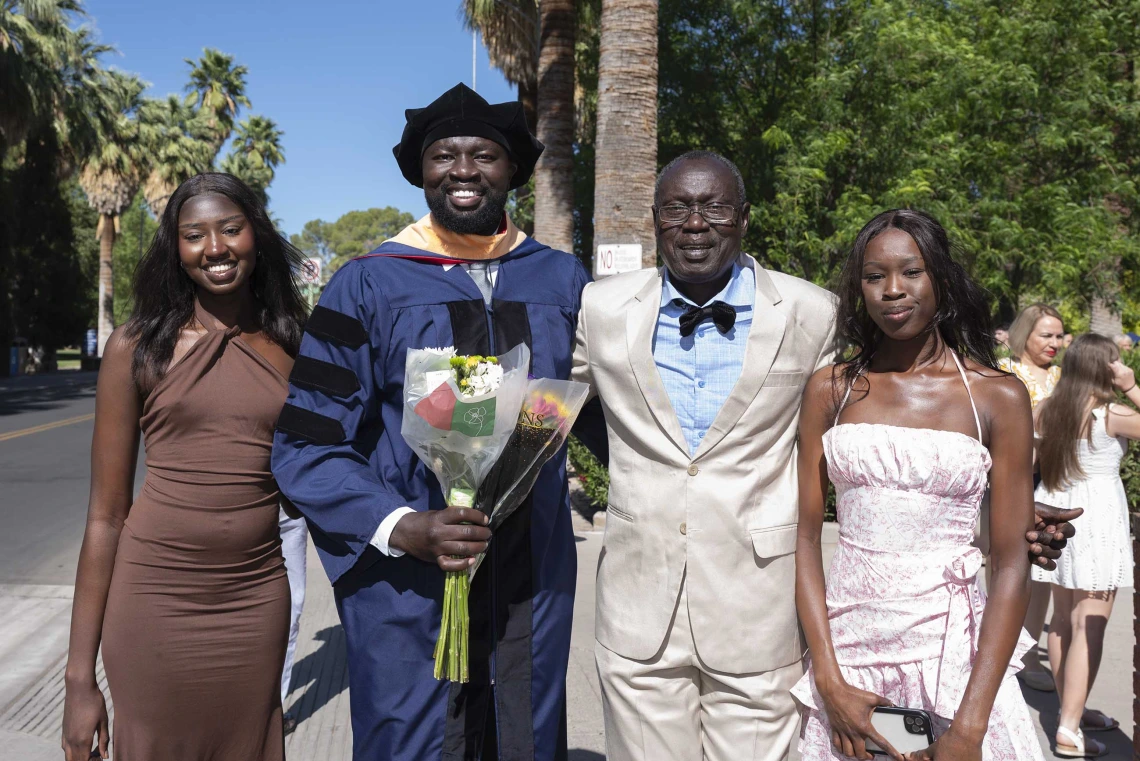 A young man in graduation regalia holds a bouquet of flowers and smiles as he stands with his two sisters and father outside. 