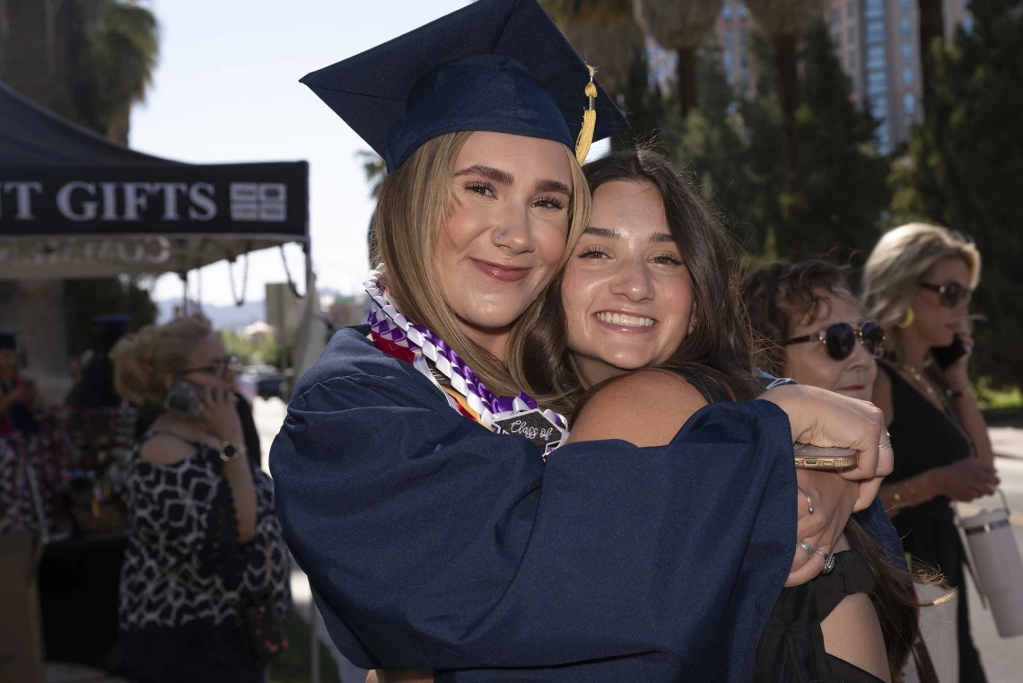 A young woman wearing a graduation cap and gown smiles as she hugs another smiling young woman. 