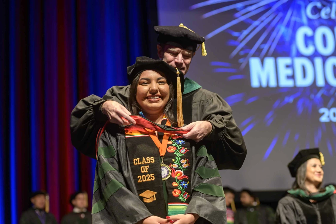 A medical student in a graduation cap and gown has a ceremonial hood placed over her shoulders by a professor in similar attire. 
