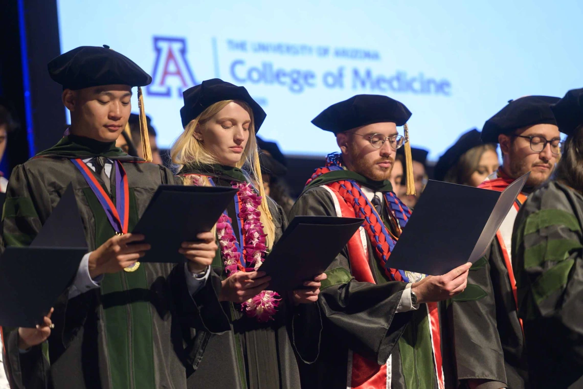 Several medical students in graduation caps and gowns read an oath from folders they are holding on a stage. 