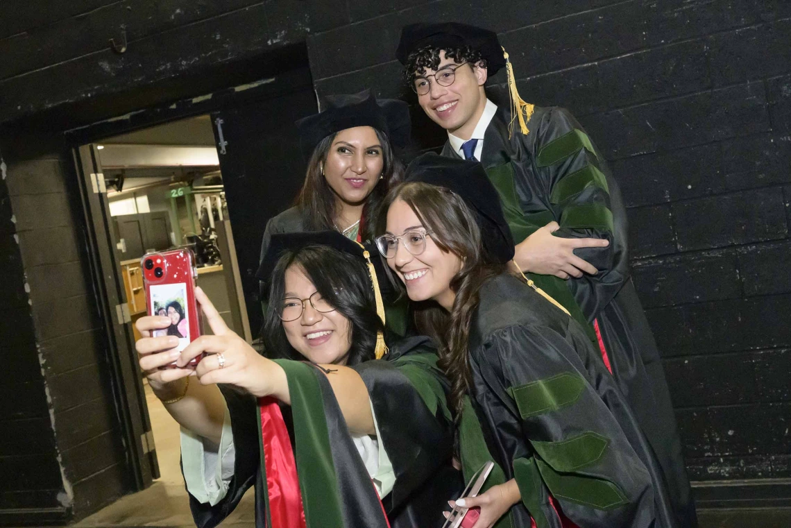 Four medical students in graduation caps and gowns bunch together to take a selfie. 