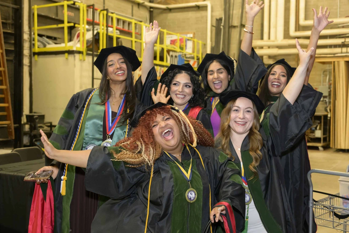 Six students dressed in graduation caps and gowns raise their hands in celebration and cheer in an industrial-looking room backstage. 