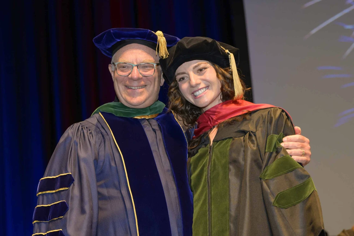 A father and daughter wearing graduation regalia smile and side hug during a graduation ceremony. 
