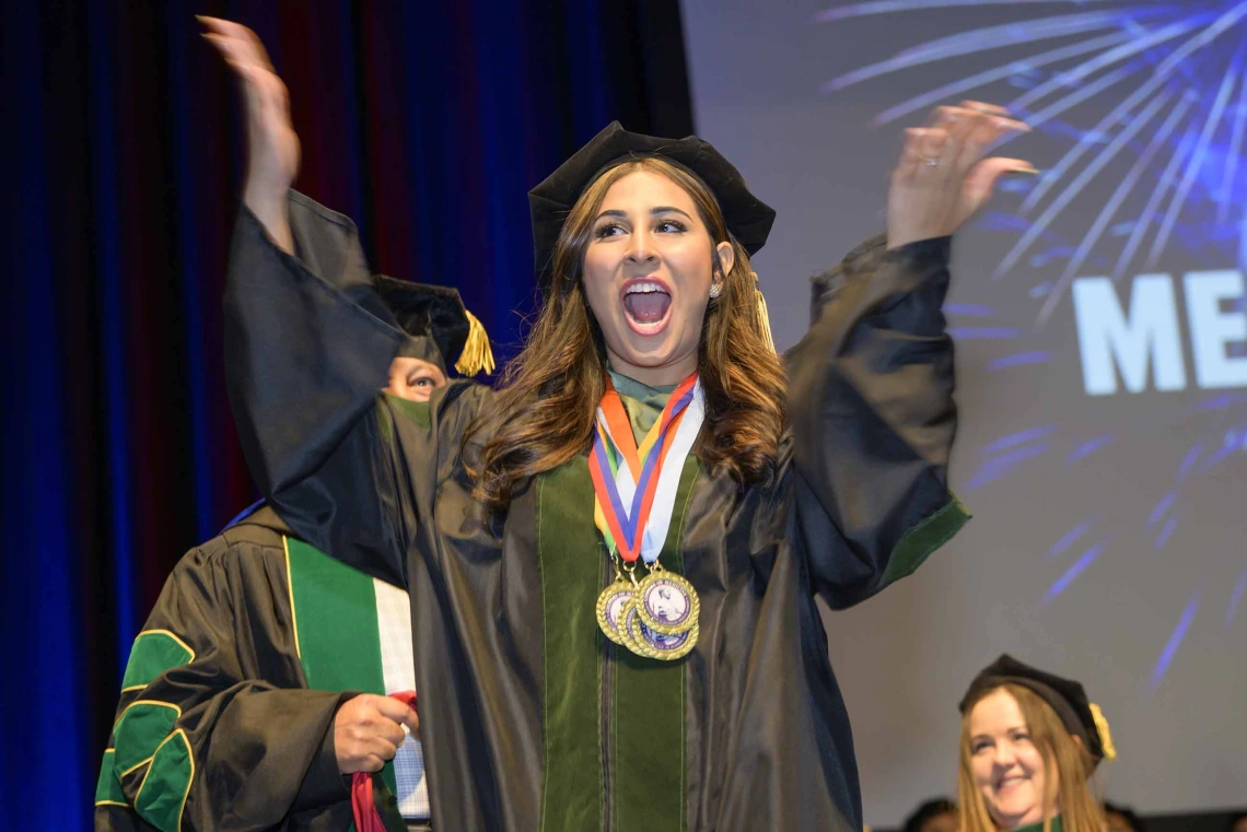 A young woman dressed in graduation regalia raises her arms in celebration with her mouth open, cheering.