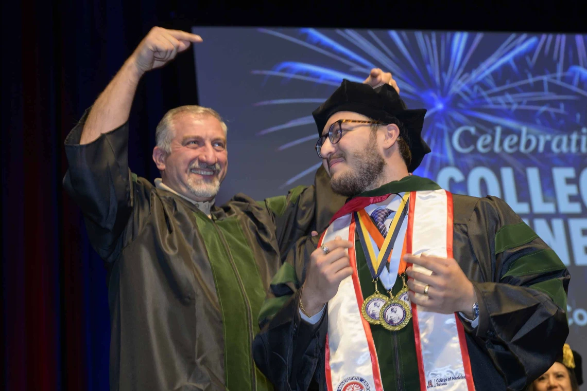 A father points in celebration at his son, both wearing graduation regalia, during a convocation ceremony. 