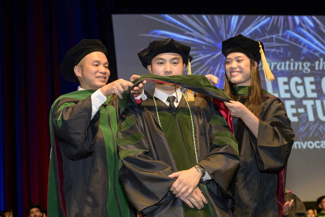 A medical student in a graduation cap and gown stands onstage as his father and sister, also in graduation regalia, place a ceremonial hood over his shoulders. 